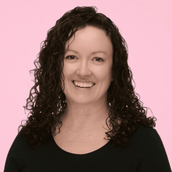 Smiling woman with long dark curly hair wearing a black top against a pink background.